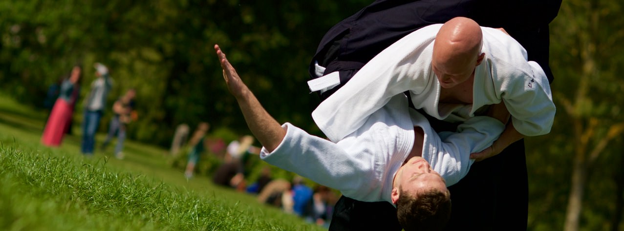 One person in a white keikogi thrown by another in a dark jacket, on the grass in a park.