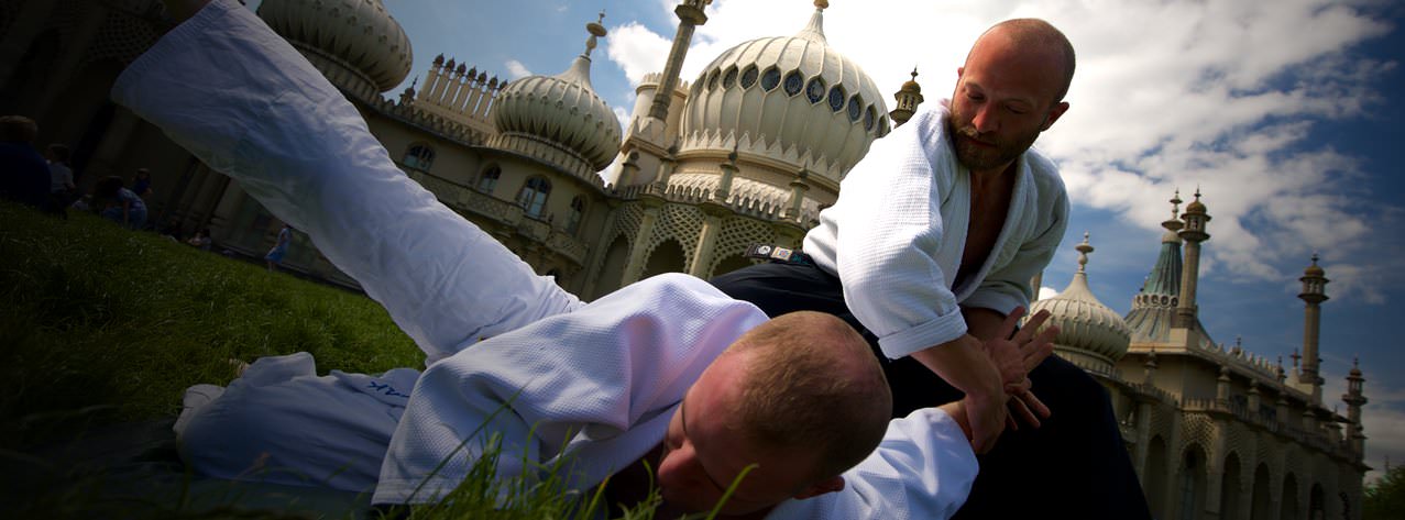 Two people in white keikogi practising a pin on the grass in front of the Royal Pavilion.