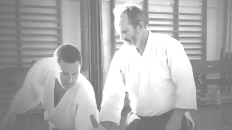 Rick Smaridge kneeling in a keikogi beside a student in a wooden training hall.